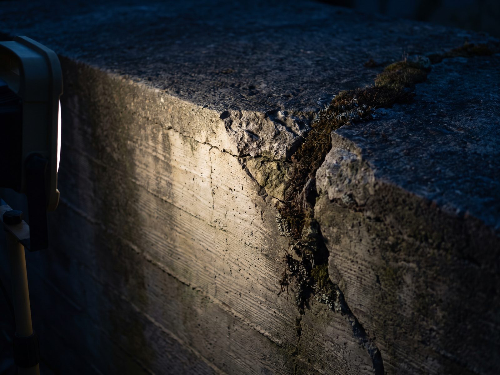 Close view of weathered cast concrete with horizontal formwork lines, a chipped edge, and a seam of moss.