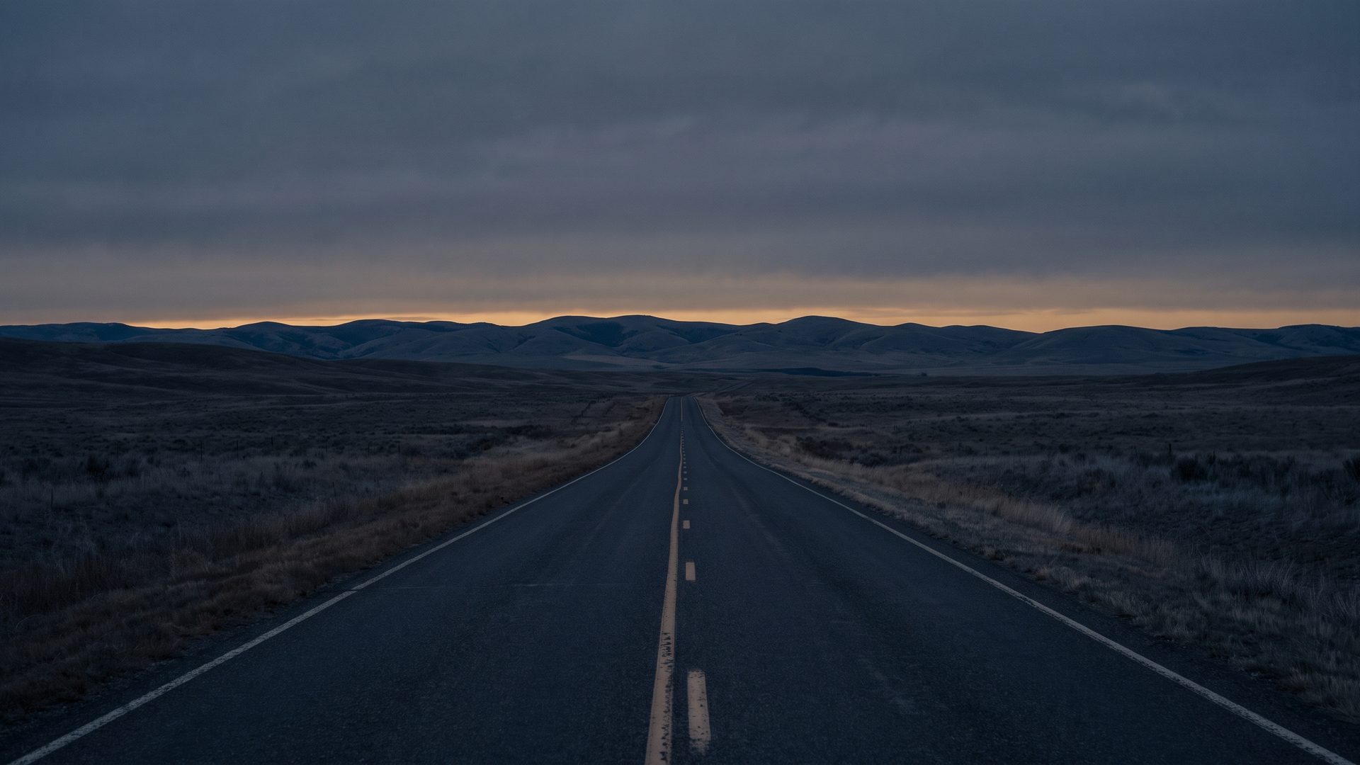 A straight empty two-lane road running toward low hills under an overcast dusk sky, with a thin amber band on the horizon.