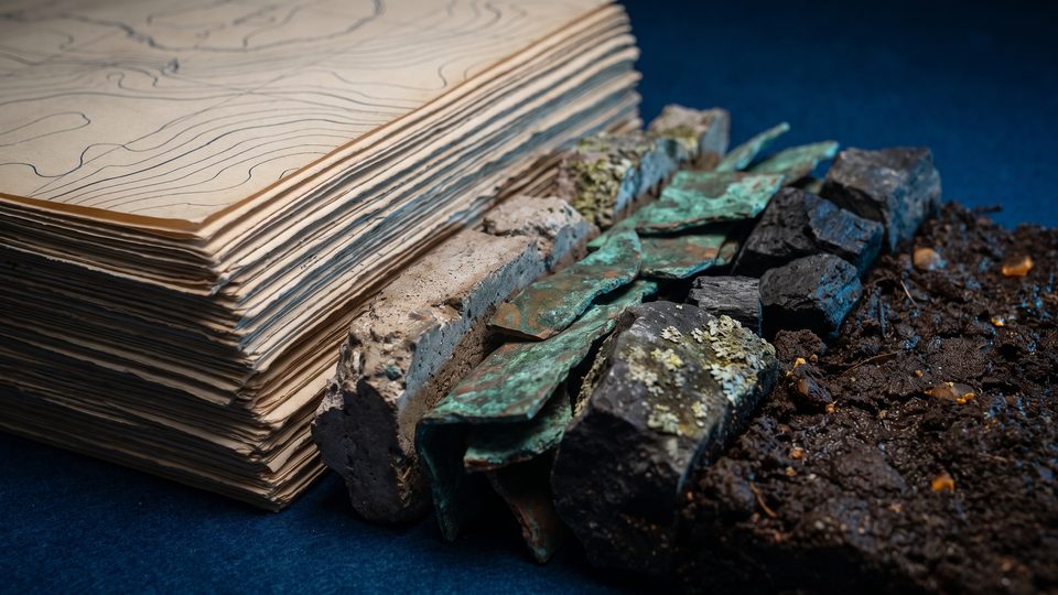 Side view of stacked paper map sheets whose lower layers become weathered concrete, oxidized copper, dark stone, and soil.